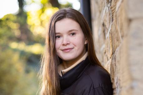 Student wearing a puffer coat leaning against a wall smiling
