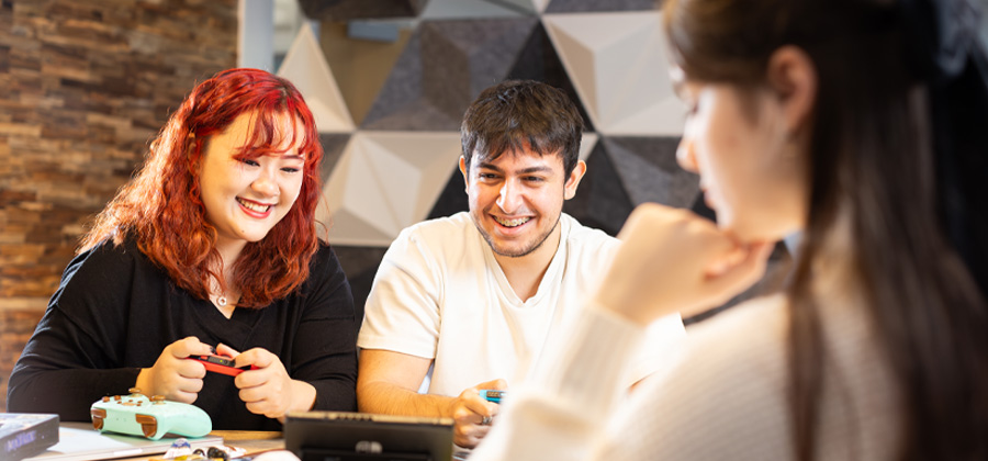Two students playing on a switch console at a table in a shared student space as another student is using art supplies in the foreground.